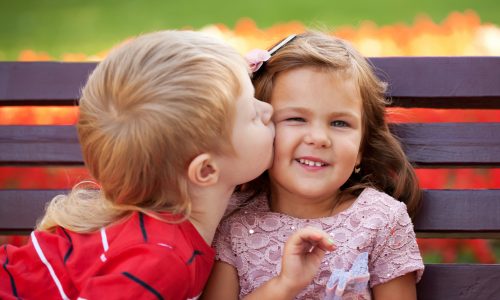A young boy kissing a smiling young girl on the cheek while sitting on a bench, demonstrating equal time and shared care.