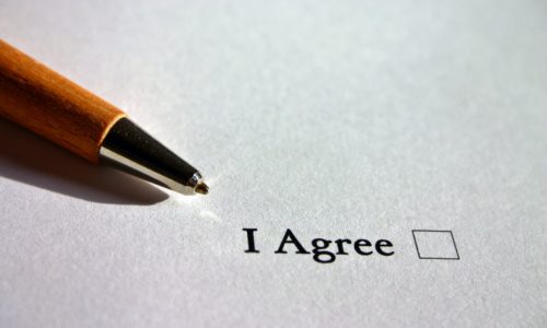 A wooden ballpoint pen near a printed document with the phrase "i agree," an unchecked checkbox, and details about a father's overseas trip.